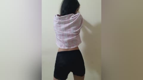 Video of a person facing away, wearing a pink striped shirt and black shorts, against a plain beige wall. The individual's back is visible, with long black hair.
