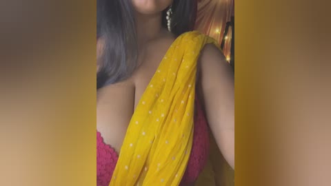 Video of a South Asian woman with long dark hair, wearing a bright yellow sari with white polka dots, draped over a red lace bra, showcasing her large breasts. She has gold earrings and a blurred background.