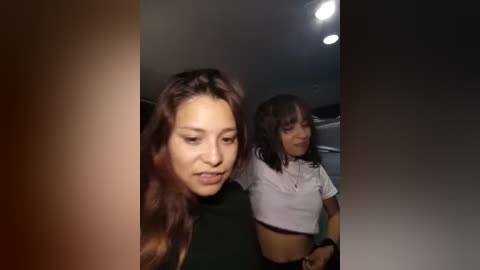 Media: Video of two young women with light brown skin, one with long wavy hair, the other with straighter hair, both wearing tight, cropped tops. They appear to be indoors with dim lighting, and the image has a grainy, low-quality filter applied.
