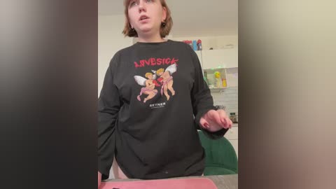 A video of a plus-sized woman in a black \"LOVE IS MAGIC\" sweatshirt with cartoon angels, standing in a kitchen.