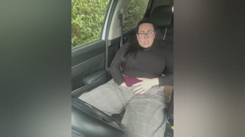 Video of a woman with glasses, medium build, black turtleneck, and gray pants, sitting in a car, holding her stomach, green foliage outside.