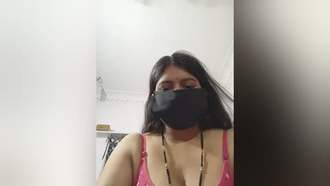 Media: Video of a woman with medium skin tone, long black hair, wearing a black face mask, pink spaghetti-strap top, and black beaded necklace, standing in a plain, white-walled room with a ceiling fan and minimal decor.