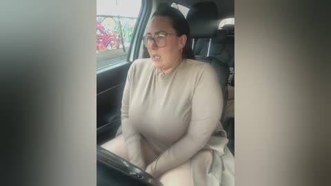 Video of a middle-aged woman with glasses, fair skin, and brown hair, wearing a beige long-sleeved top, sitting in a car with black leather seats.