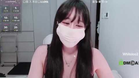 A video of a young Asian woman with long black hair, wearing a white face mask, a black tank top, and a necklace, sitting indoors with a digital thermometer on the wall.