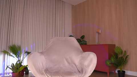 A video of a modern room featuring a cream-colored, fabric-covered chair, light wooden walls, a red cabinet, green plants, and beige curtains.