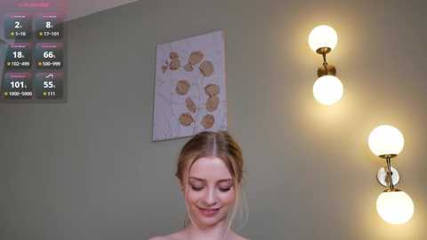 Video of a smiling young woman with light skin and blonde hair in a simple room with beige walls, a floral art piece, and two wall-mounted lamps.