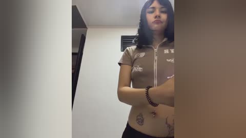 A video of a young woman with medium skin tone, dark hair, and a slim physique, wearing a brown crop top and black pants. She has tattoos on her abdomen and arms. The background is a white wall with a vent.