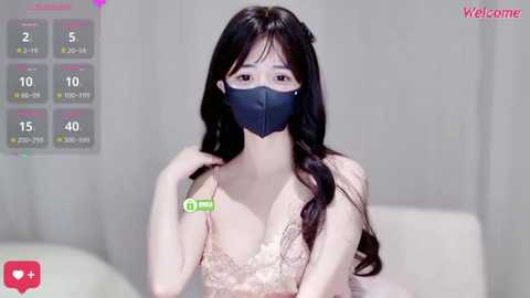 Media: Video of an East Asian woman with long black hair, fair skin, wearing a black face mask, beige lace bra, and white top, sitting on a white couch. Background shows a simple, modern room.