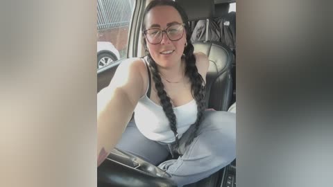 Video of a smiling woman with glasses and long black braided hair, wearing a white tank top and blue jeans, seated in a car, with a blurred background.