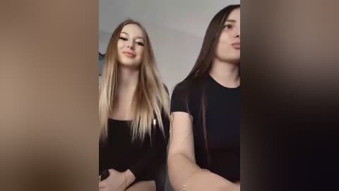 Video of two young women with long, straight hair, smiling. One has blonde hair, the other dark brown hair. They wear black tops.