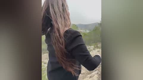 Video of a person with long, wavy brown hair in a black jacket, standing outdoors on a grassy hillside with distant, foggy mountains in the background.