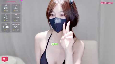 A video of an East Asian woman with short, straight brown hair, wearing a black face mask, black halter top, and a green name tag, giving a peace sign. The background is blurred.
