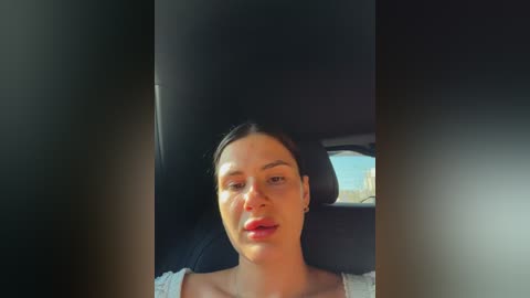 Video of a woman with dark hair and fair skin, wearing a white top, sitting in a car with a dark interior, sunlight filtering through the window, creating a warm, natural ambiance.