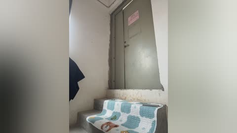 Video of a narrow, dimly-lit corridor with peeling white walls, a gray door, and a torn blue curtain. A white blanket with blue and pink patterns is draped over a metal bench.