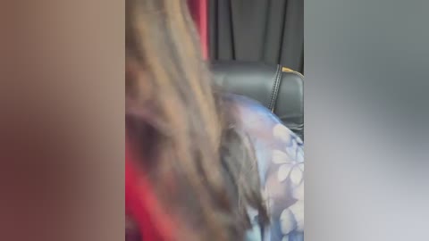 Video of a person with shoulder-length blonde hair, wearing a blue floral-patterned shirt, seated on a black leather chair with a red curtain in the background.