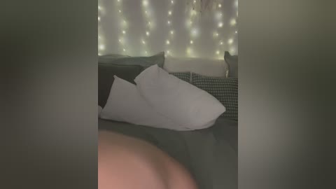 A video of a dimly-lit bedroom with a cozy, dark green bedspread, two white pillows, and soft, warm fairy lights on a white wall. The focus is on the pillows, creating a serene, intimate atmosphere.