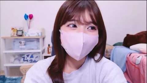 Video of an Asian woman with long brown hair, wearing a white mask, light blue shirt, and sitting in a cluttered, brightly lit room with shelves, toys, and a bed in the background.