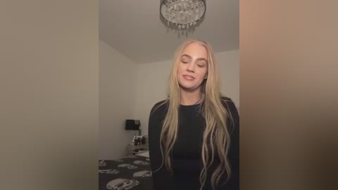 Video of a young, smiling, fair-skinned woman with long, wavy blonde hair in a black sweater. She stands in a beige room with a chandelier and a bed with skull-patterned bedding in the background.