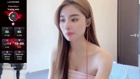 A video of an Asian woman with long brown hair, wearing a pink strapless dress, seated indoors, listening to music on red earbuds. The background shows a white wall and a digital screen displaying music controls.