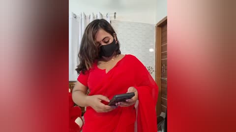 Video of a woman in a bright red sari, mask, and glasses, holding a smartphone, in a modern, softly lit room with white walls and wooden furniture.
