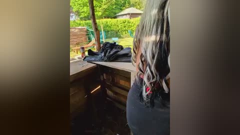 Video of a person with long, wavy, blonde hair, wearing a black shirt and dark pants, standing in a dimly lit kitchen with wooden cabinets and a glass door leading to a sunny backyard.