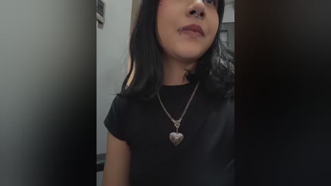 Video of a young woman with medium-dark skin, black hair, and a septum piercing, wearing a black top and heart-shaped necklace, standing in a dimly lit room.
